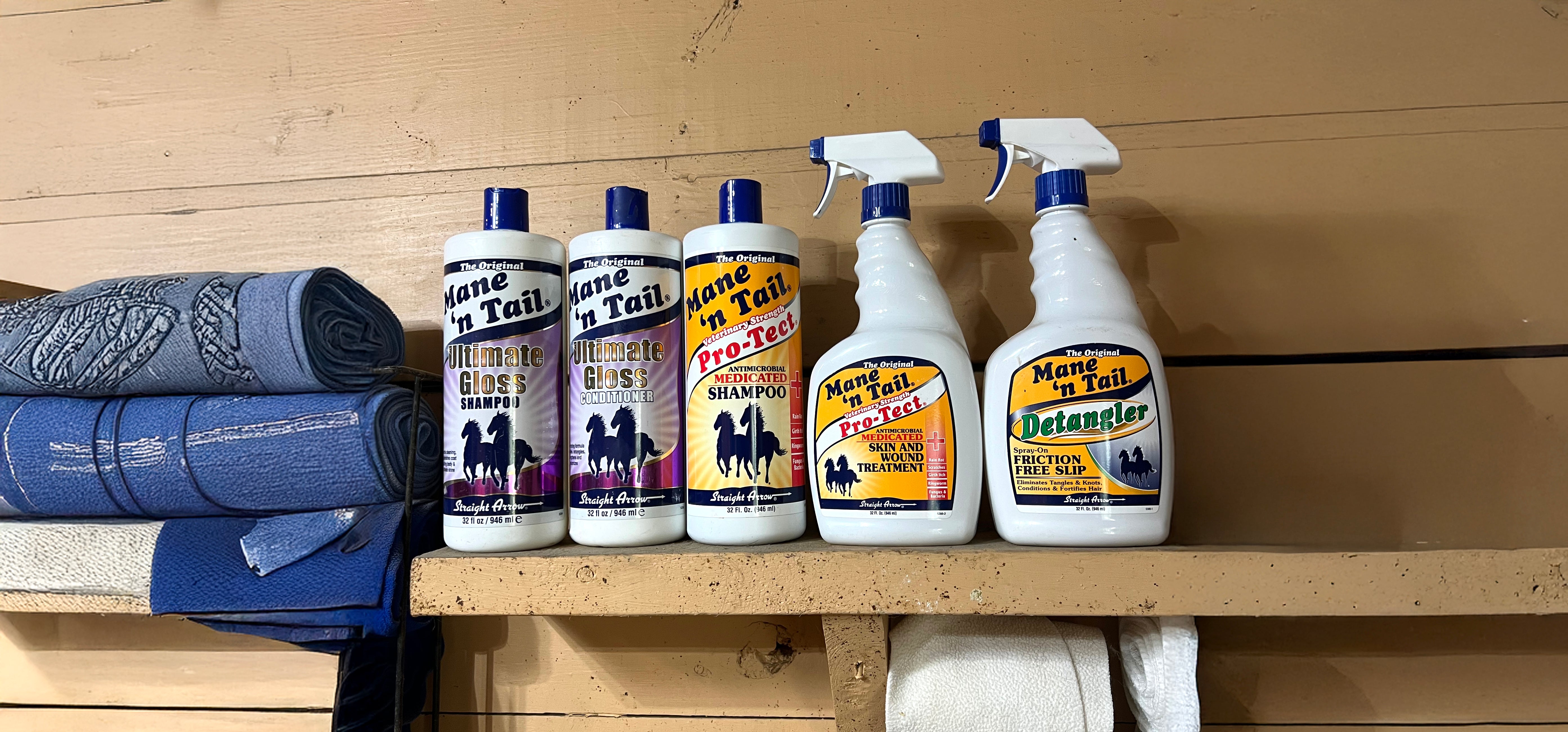 A tan shelf holding some Mane 'n Tail products, from left to right. Mane 'n Tail Ultimate Gloss Shampoo, Mane 'n Tail Conditioner, Mane 'n Tail Pro-Tect Antimicrobial Medicated Shampoo, Mane 'n Tail Antimicrobial Medicated Pro-Tect Spray, Mane 'n Tail Detangler Spray. Next to the products on the left are some blue towels neatly folded