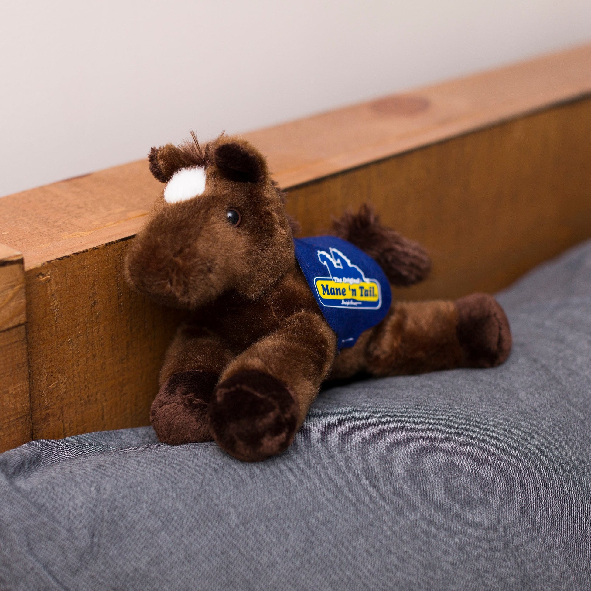 Small Chestnut horse stuffed animal wearing a royal blue branded mane 'n tail saddle. Standing on a cushion