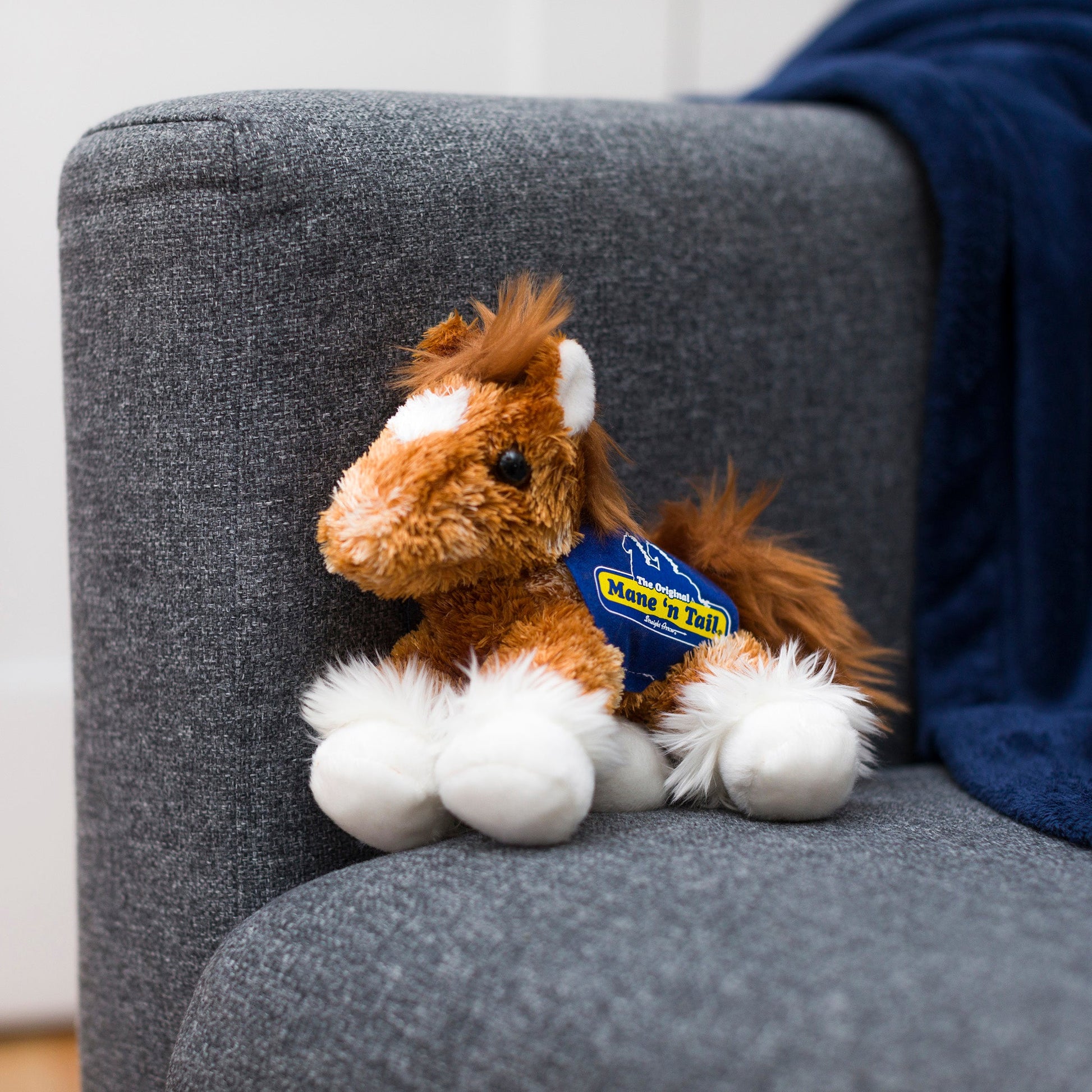 Small orange Clydes horse stuffed animal wearing a royal blue branded mane 'n tail saddle. Standing on a couch