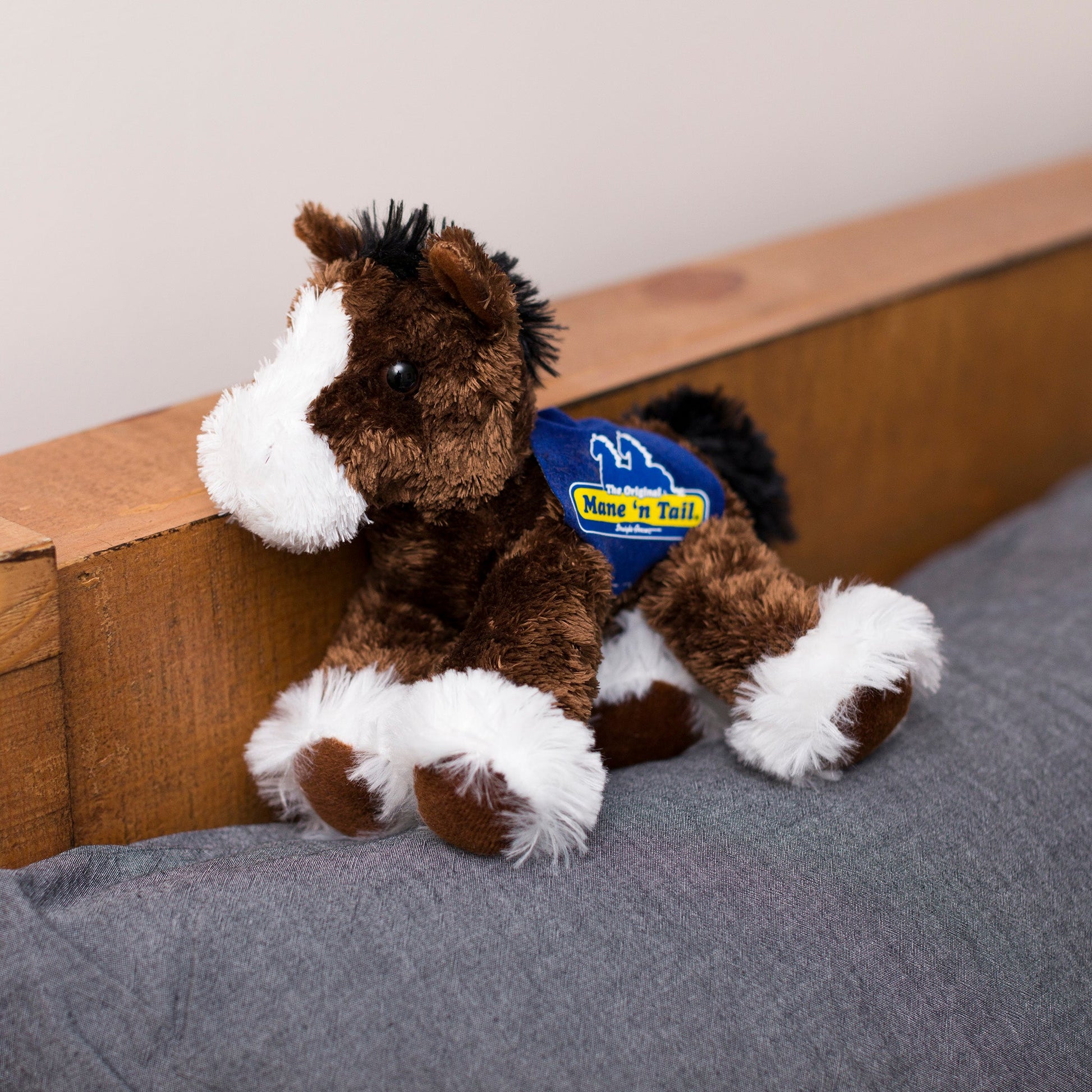 Small brown Clydes horse stuffed animal wearing a royal blue branded mane 'n tail saddle. Standing on a cushion