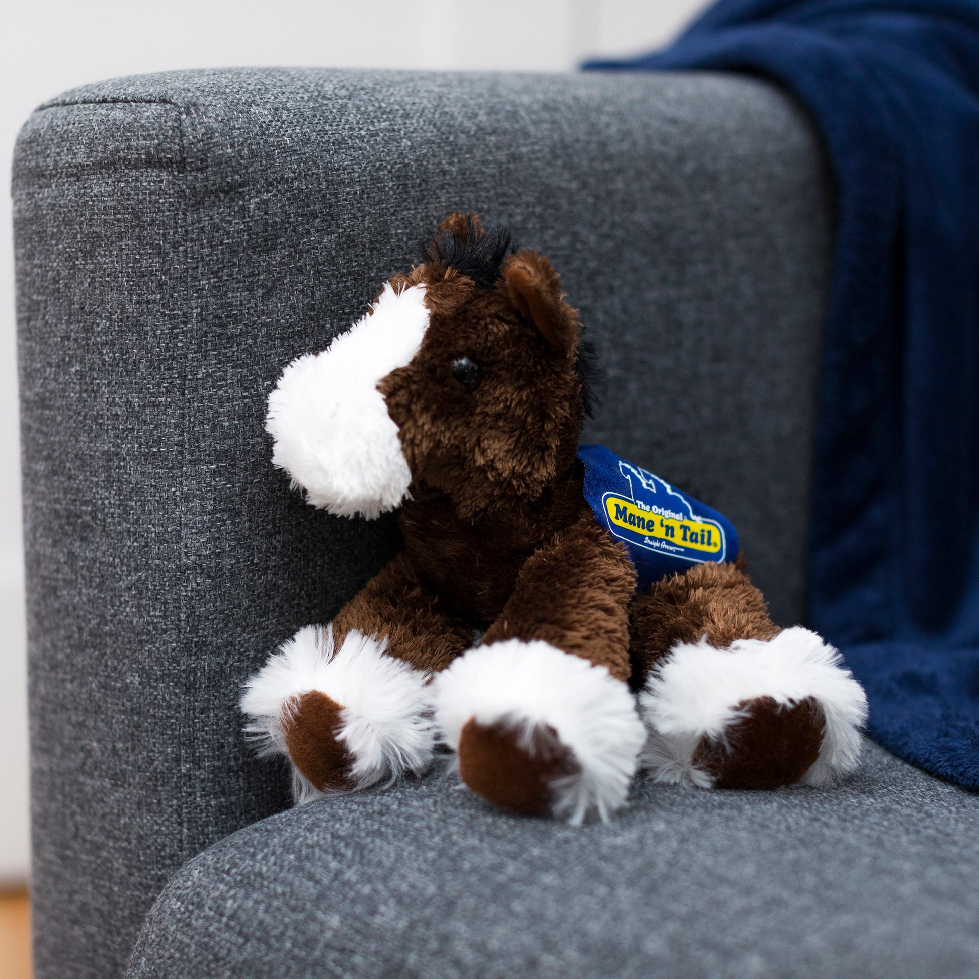 Small brown Clydes horse stuffed animal wearing a royal blue branded mane 'n tail saddle. Standing on a couch