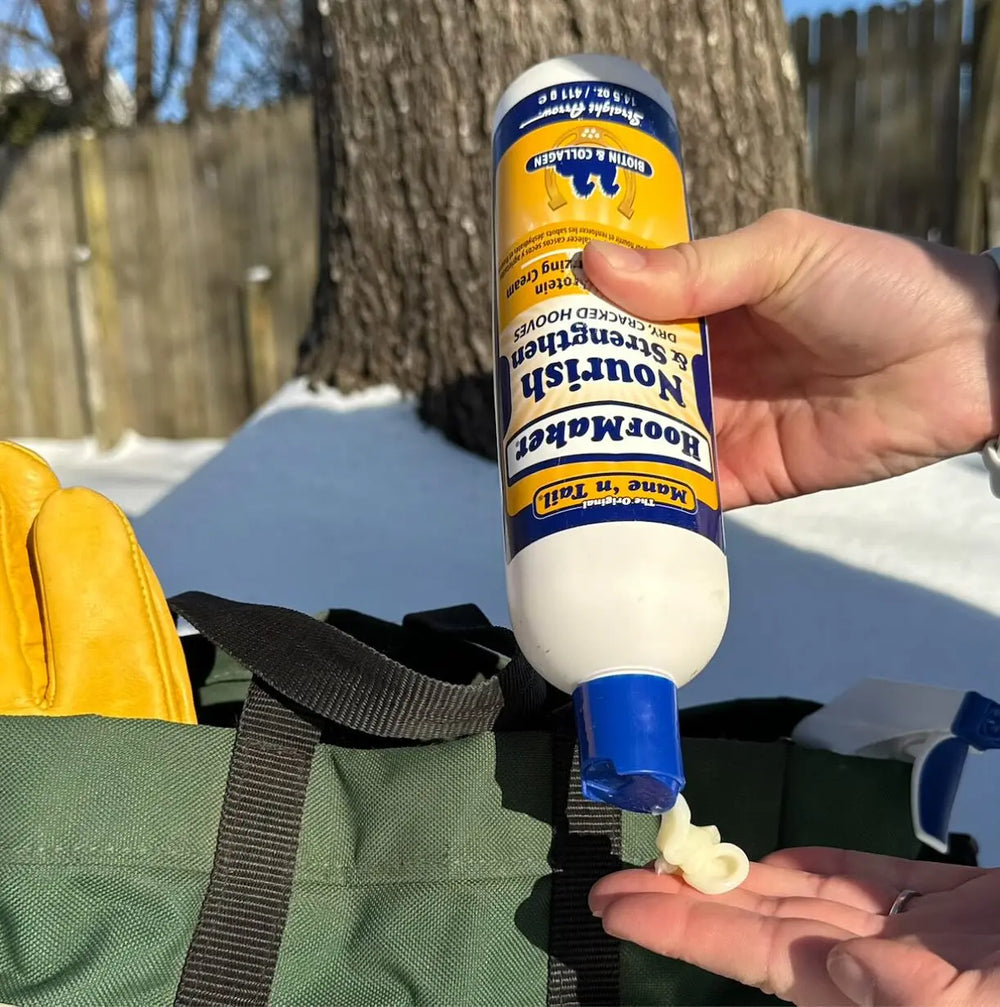 A person applying 14.5 Ounce Mane 'n Tail Hoofmaker. In the background there is some snow and a tree, closer there is a green tack bag