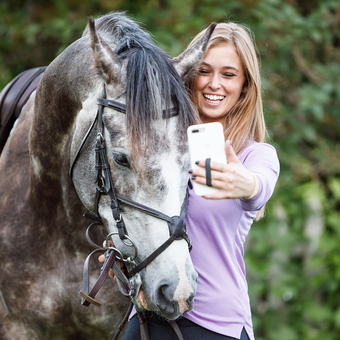 A woman holding a phone outward. The women is standing next to a horse